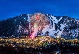 A vibrant display of fireworks illuminates the night sky above the snow-covered mountains, casting a colorful glow over the twinkling lights of the town below. The majestic peaks serve as a dramatic backdrop, highlighting the festive atmosphere.