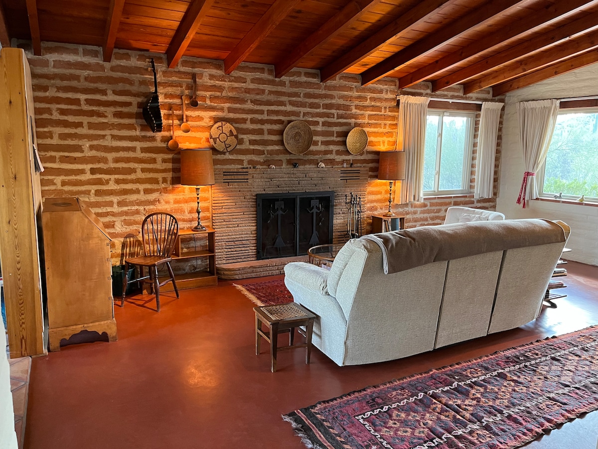 A spacious living area features a cozy sofa facing a rustic fireplace, with wooden accents and artfully arranged decor on the brick walls. Natural light enters through large windows, illuminating the warm tones of the floor and the nearby vintage items.