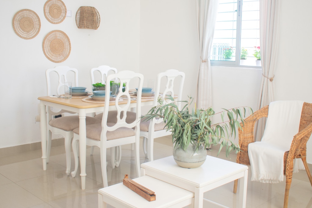 A dining area is presented with a white wooden table surrounded by six light-colored chairs. Woven wall decor enhances the space, while a potted plant occupies a coffee table, accompanied by a small decorative item. Natural light filters through the nearby window.