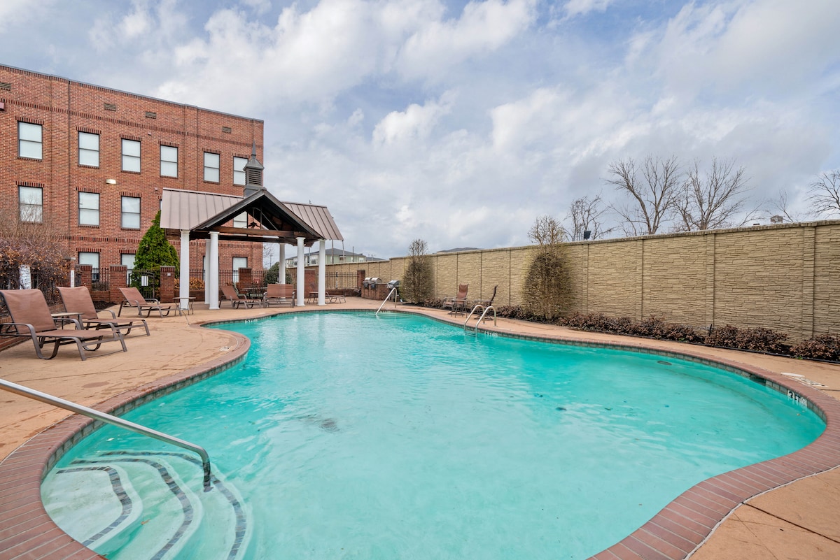 A shared swimming pool is surrounded by well-maintained landscaping and features both ladder access and a gently sloping entry. Lounge chairs are positioned around the pool, while a shaded pavilion with a roof provides a space for relaxation. The area is designed for warm-weather enjoyment.