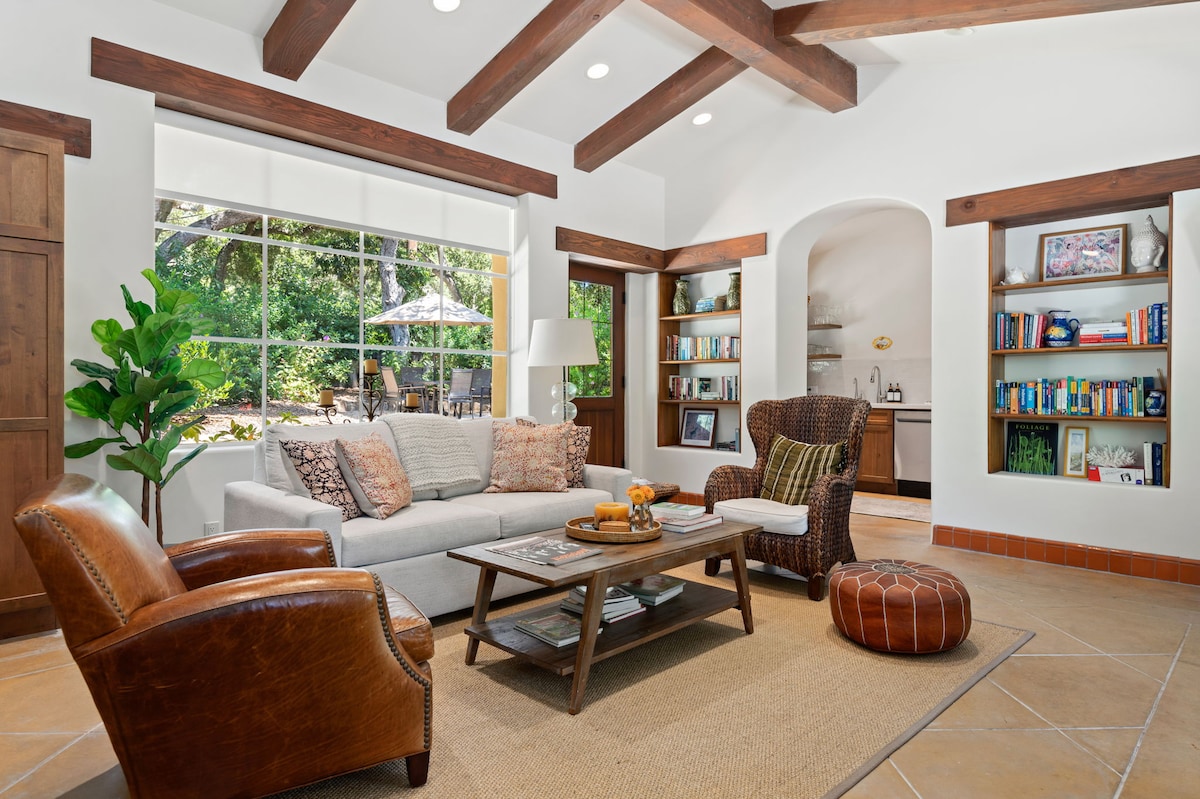 A cozy living room is highlighted by a spacious sofa and armchairs, with warm wood beams above. Natural light fills the space through large windows overlooking a private outdoor area. Bookshelves line the walls, filled with various titles, and a coffee table rests on a textured area rug.