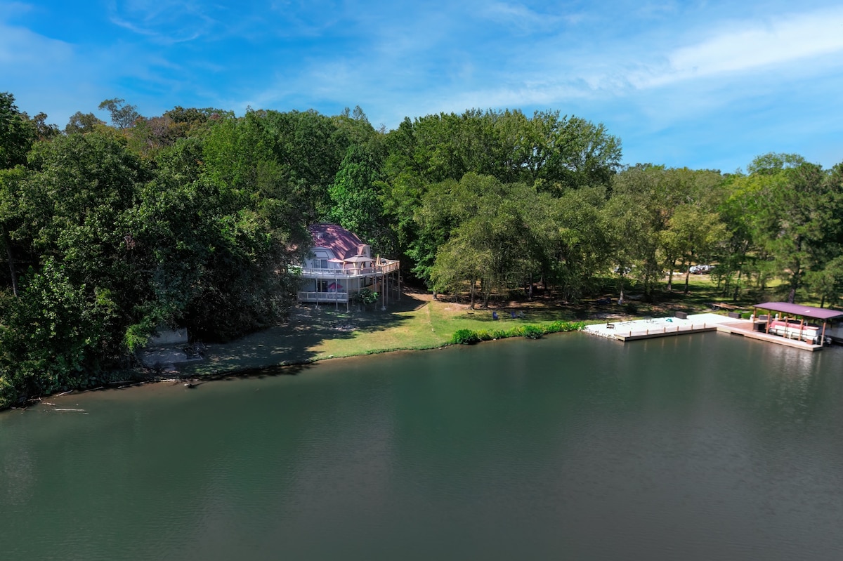 A geometric home is nestled among lush trees on the lakeshore, surrounded by a spacious yard that gently slopes towards the water. The calm lake reflects the sky, and a private dock is visible nearby, providing access for boats.