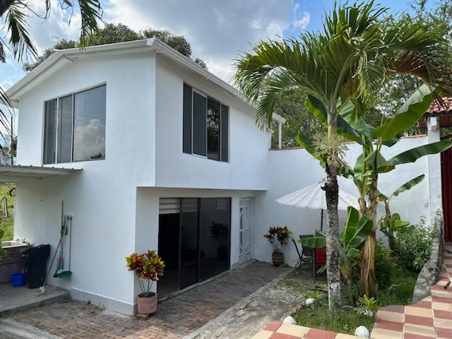 The exterior of the guest house features a two-story white building with large glass windows allowing natural light. Lush greenery, including palm and banana trees, surrounds the space. A patio umbrella is positioned near vibrant plants, creating a welcoming entryway.