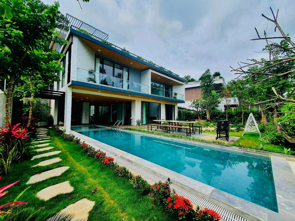 A modern villa is depicted with a glass facade overlooking a clear swimming pool. Lush greenery and flower beds line the pathway leading to the pool area. A spacious outdoor dining table is set on the patio, surrounded by well-maintained gardens.