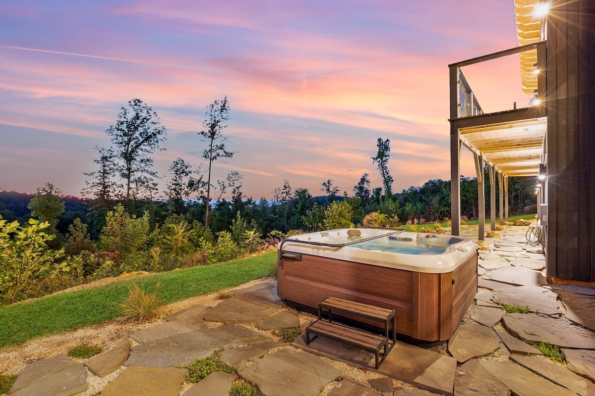 A hot tub is set on a stone patio, surrounded by lush greenery and offering panoramic views of the landscape. The sky is painted in soft pink and orange hues during sunset, enhancing the serene environment.