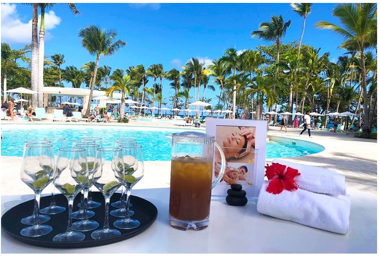 A refreshing scene is presented with a tray of champagne flutes arranged beside a pitcher of iced tea. A framed photo and neatly rolled white towels accompany the items on a table, with a sparkling pool and palm trees softly blurred in the background.