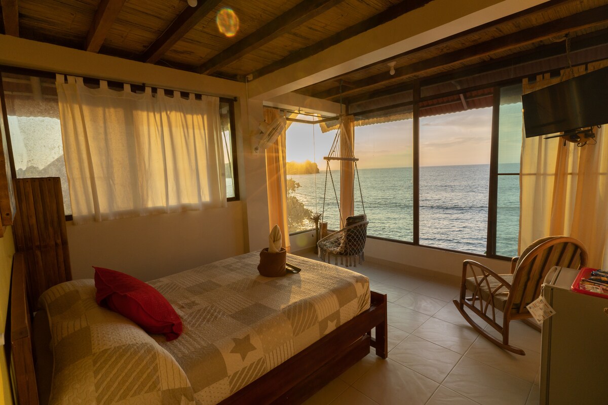 A serene bedroom features a spacious bed dressed in a neutral-toned quilt. Floor-to-ceiling windows provide a view of the ocean, while soft curtains gently diffuse the light. A rocking chair and swing chair are positioned to enjoy the coastal scenery.