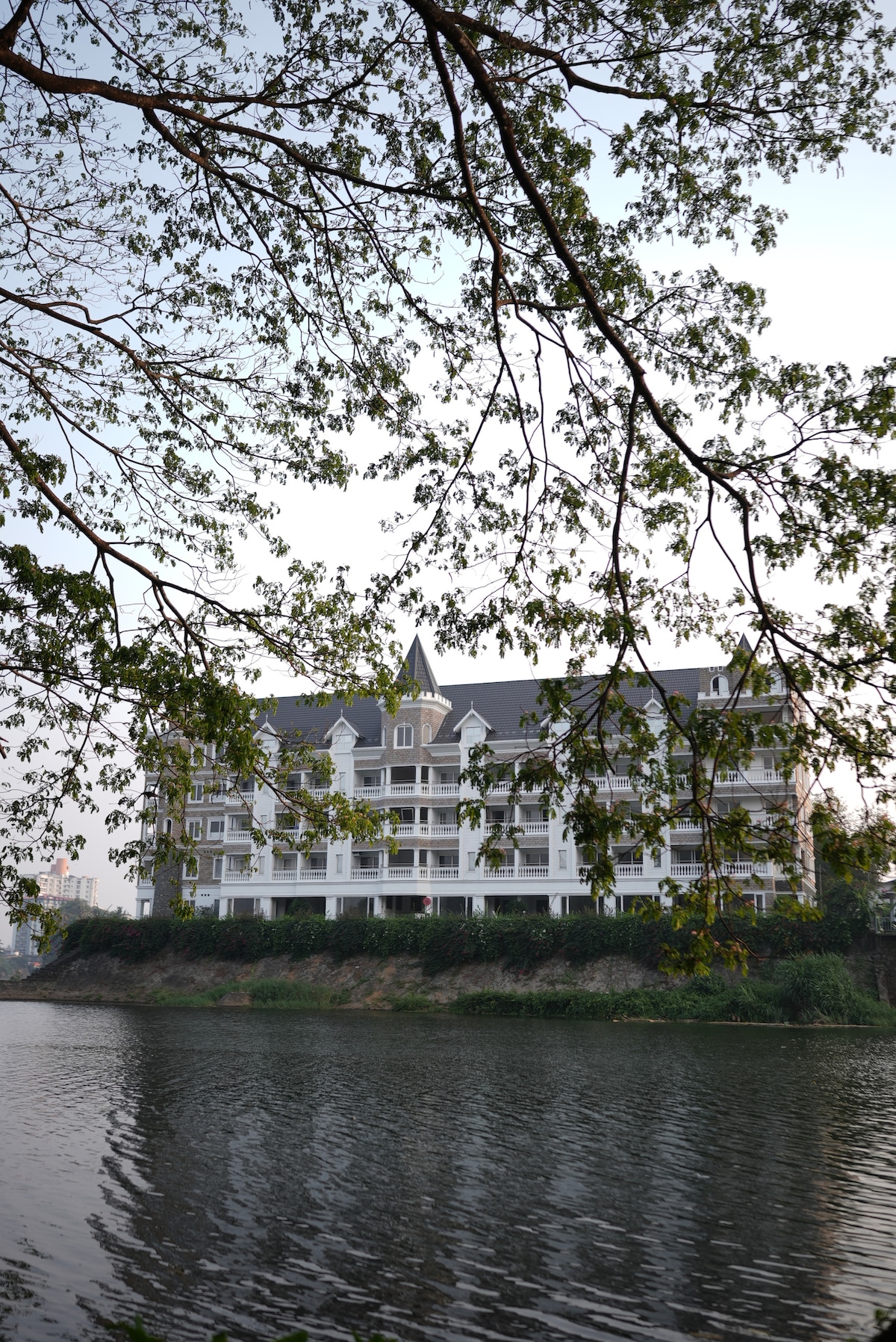 A modern duplex apartment is reflected in the calm waters of the Periyar River. Framed by branches and leaves, the building showcases multiple levels with balconies, blending in with the scenic environment. The serene atmosphere invites relaxation and offers picturesque views from the riverbank.