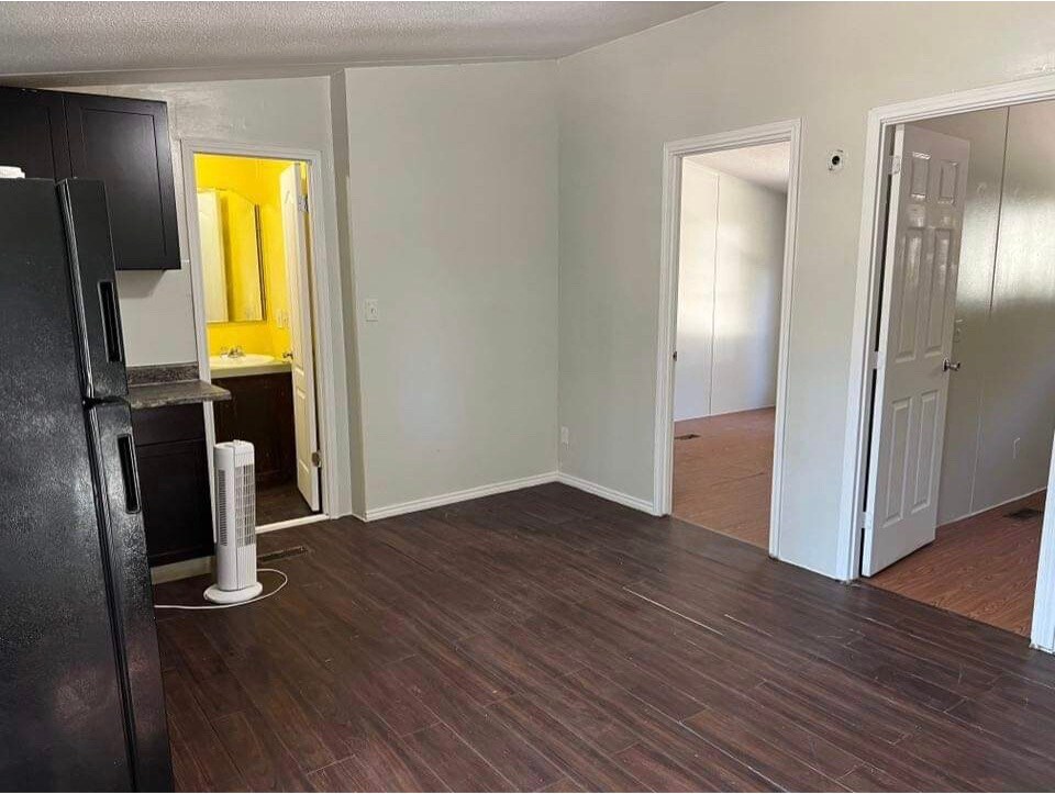An open kitchenette space features dark cabinetry and a countertop, with a visible black refrigerator. A doorway leads to a bathroom, while another door opens to a separate room. Light brown flooring adds warmth to the area, creating an inviting environment.