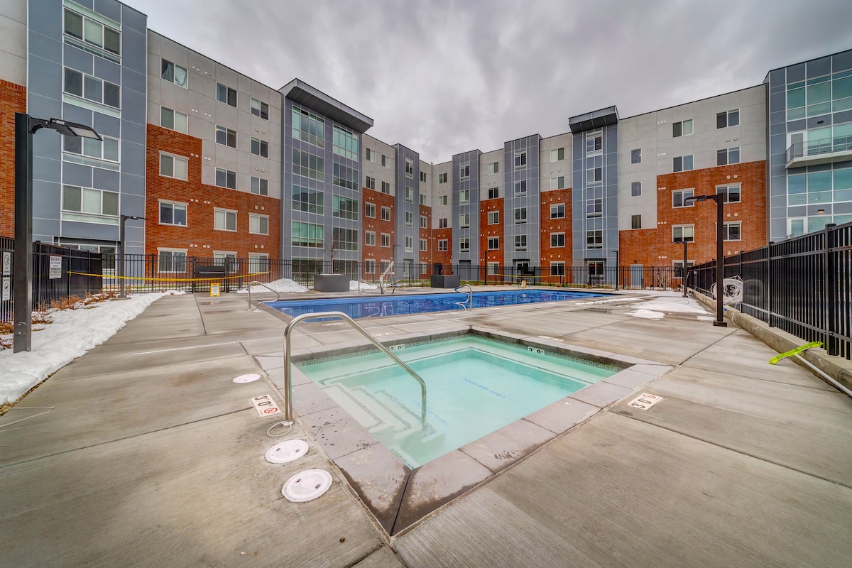 A modern, outdoor pool area is visible, featuring clear water in a large swimming pool and an adjacent hot tub. The surrounding pavement is smooth, and contemporary apartment buildings stand in the background, displaying a mix of brick and grey exteriors.