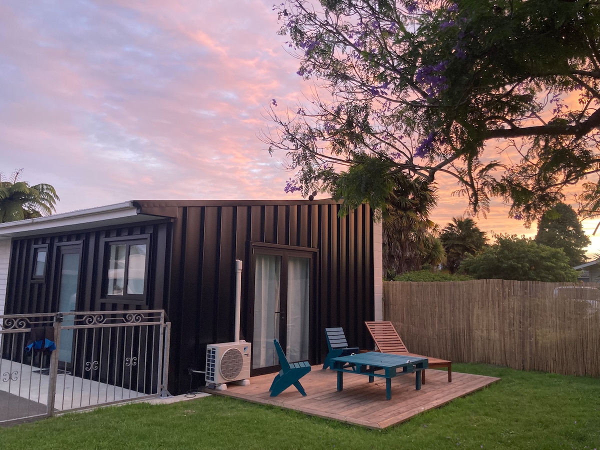 A modern studio with a sleek black exterior is illustrated against a vibrant sunset sky. Outdoor seating is arranged on a wooden deck, complemented by green grass. The entrance features large windows, allowing natural light to fill the space.