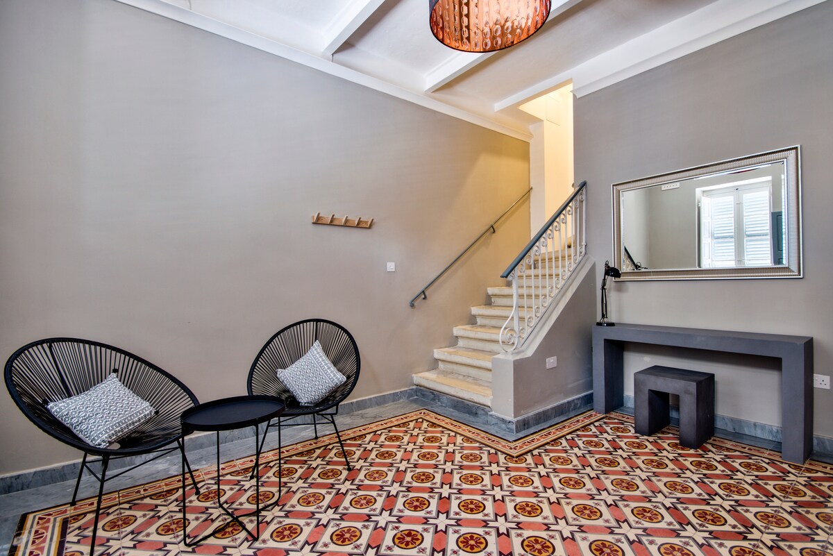 A spacious entrance hall is framed by unique Maltese tile flooring, showcasing a neutral gray wall. Two black chairs with cushions are positioned beside a small table, creating a welcoming seating area. A staircase leads to the upper levels, with a mirror reflecting natural light.