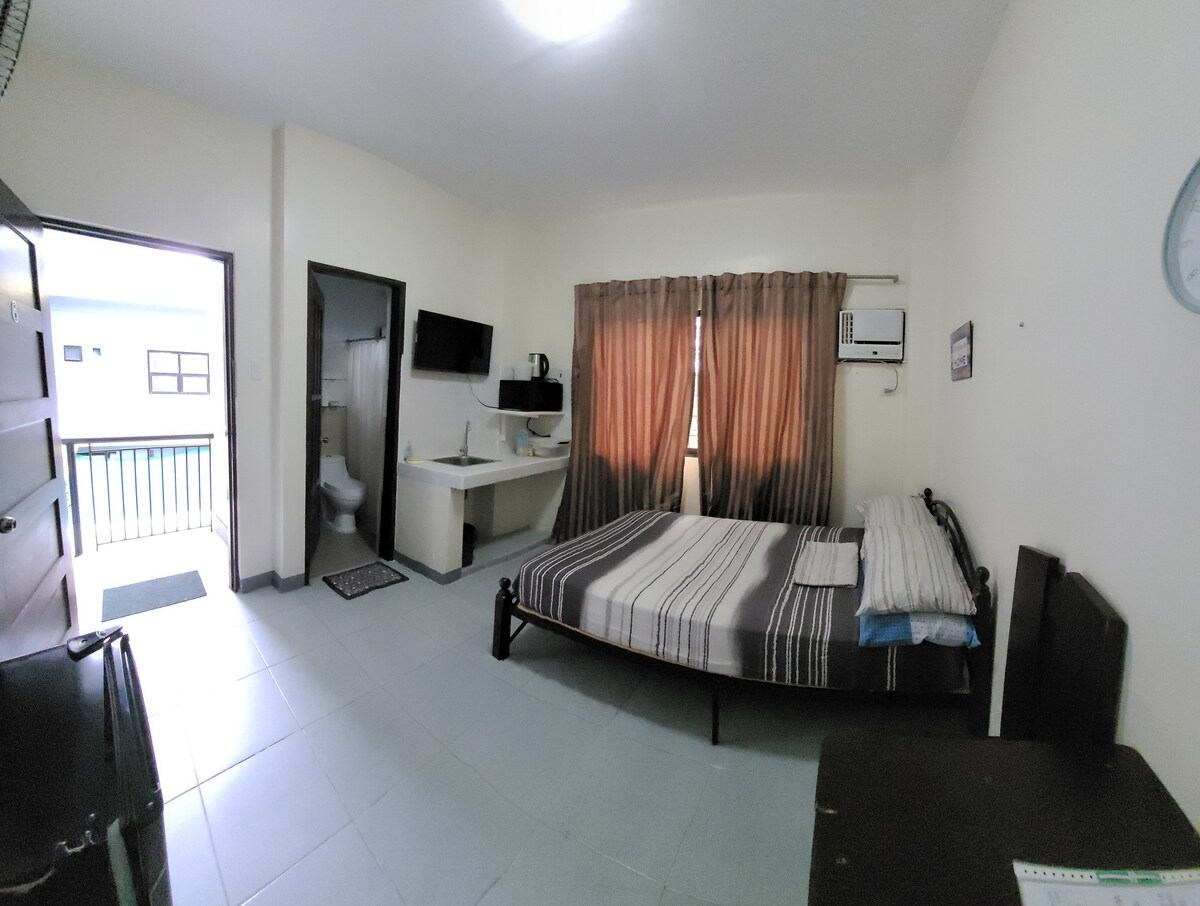 A well-lit studio space features a spacious bed with striped bedding, a simple desk area with a monitor, and a wall-mounted smart TV. Large windows with brown curtains allow natural light to filter in, while a private bathroom door is visible in the background.