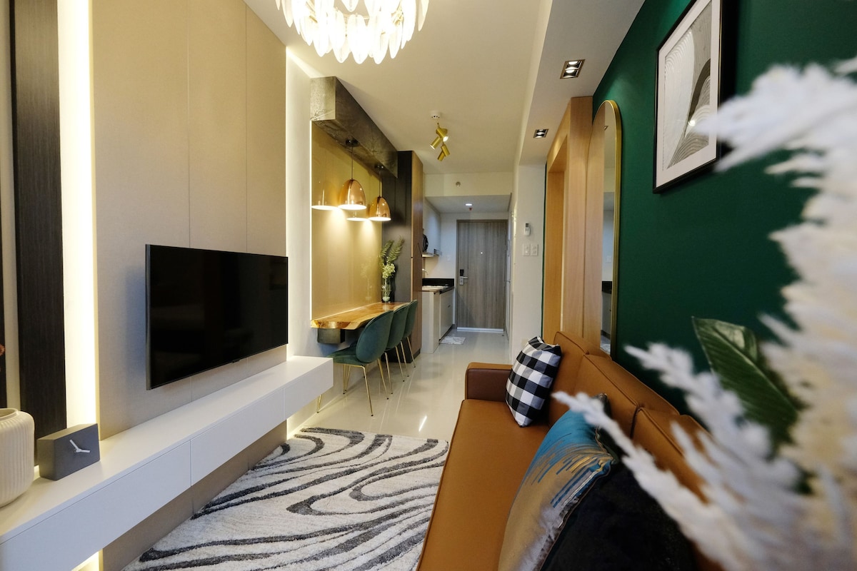 A modern living area is shown, featuring a sleek television mounted on a wall. A cozy leather couch is positioned across from the TV, complemented by a textured area rug. A dining table with colorful chairs is visible in the background, alongside softly illuminated shelving.