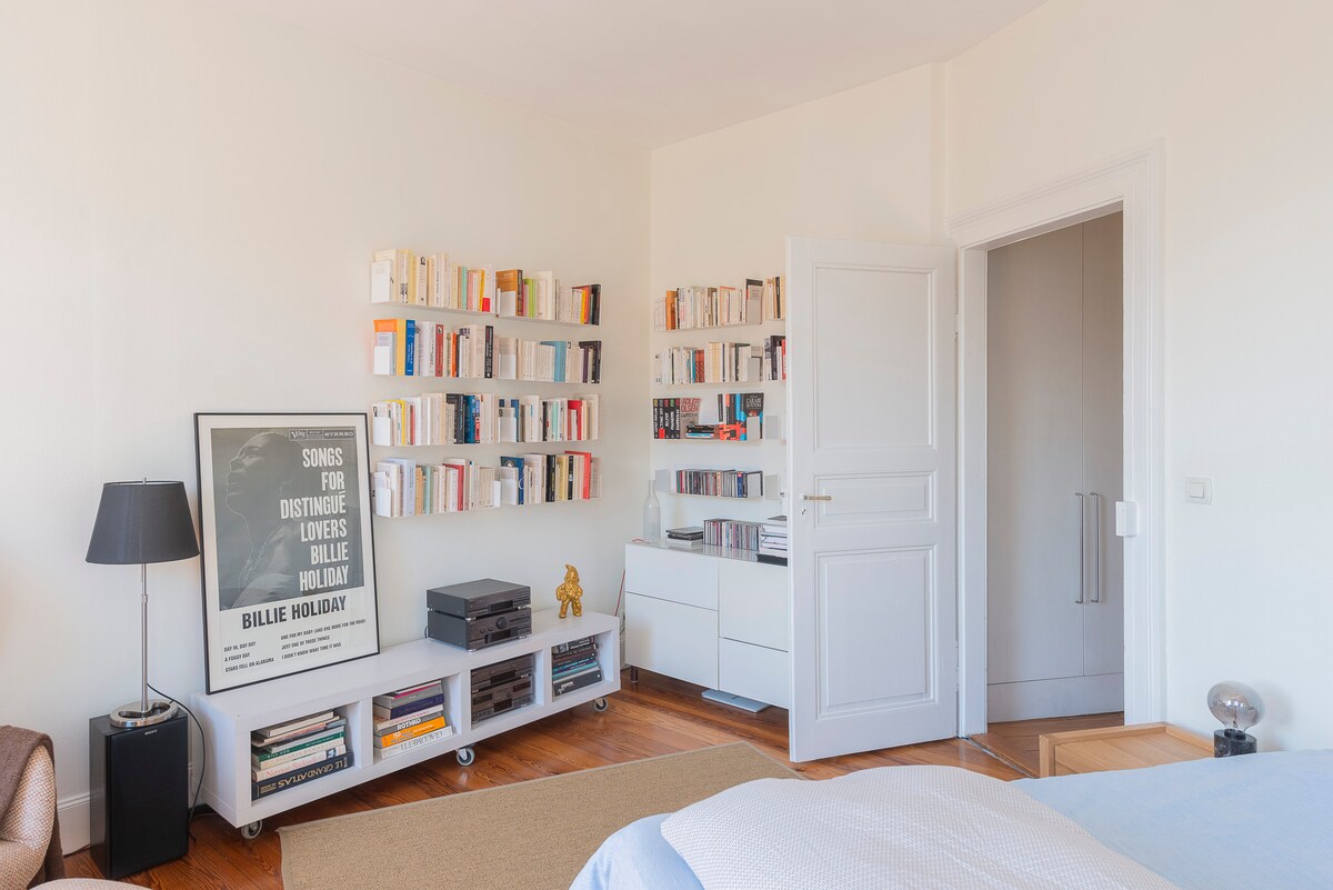 A well-lit bedroom features a cozy bed complemented by a reading area with a sleek armchair. A bookshelf filled with various books is positioned against the wall, while a stylish lamp sits nearby. A modern white cabinet is also visible, offering additional storage.