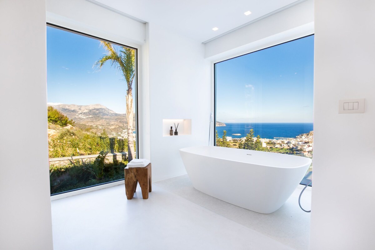 A modern bathroom features a freestanding bathtub positioned before large windows offering panoramic views of the sea and surrounding hills. A small wooden stool is placed nearby, contributing to the minimalist aesthetics of the space, which is illuminated by natural light.