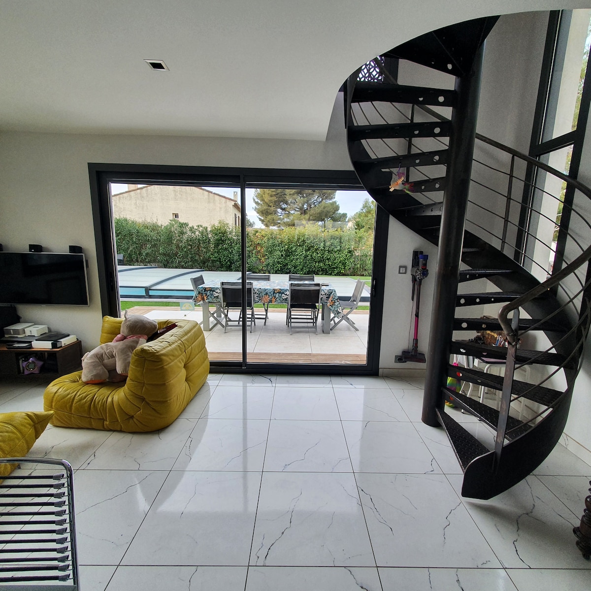 A modern living space features a spiral staircase that leads to the upper level. Large glass doors open to a terrace, revealing outdoor dining furniture surrounded by greenery. The room is furnished with a yellow lounge chair, enhancing the bright and open atmosphere.