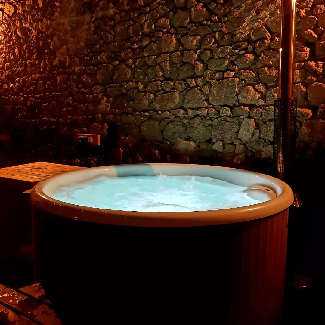 A round, wooden hot tub is set against a textured stone wall, illuminated by warm lighting. The water inside is bubbling and appears inviting, creating a relaxing ambiance in this private outdoor space.