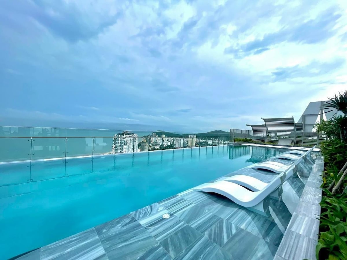 An outdoor infinity pool is seen, featuring sleek lounging chairs partially submerged in water. The backdrop includes a panoramic view of the city skyline and distant hills, set under a cloudy sky that enhances the tranquil atmosphere.