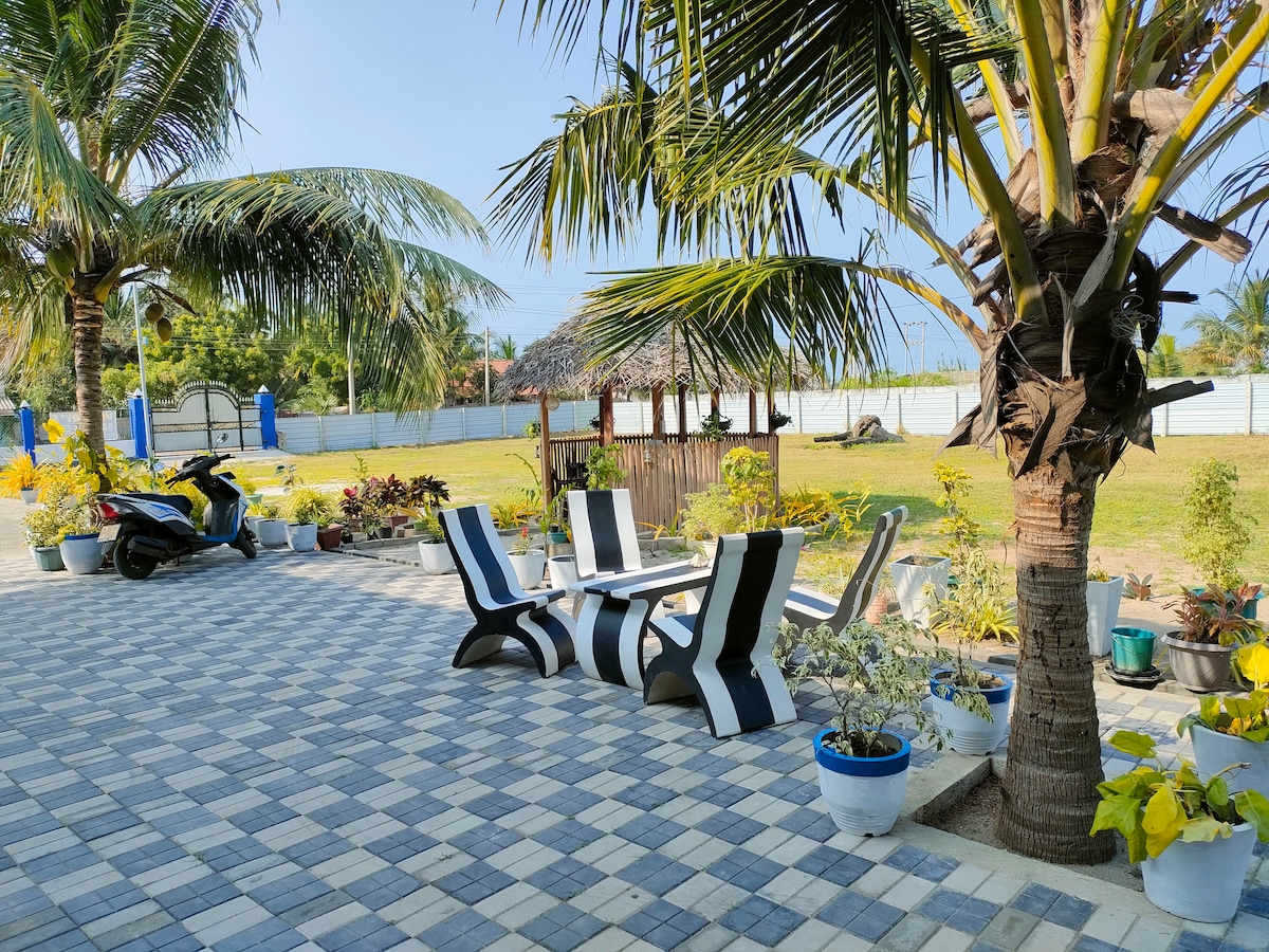 A spacious outdoor area features patterned tiled flooring with several lounge chairs arranged under palm trees. Potted plants add greenery around the space, while a gazebo is visible in the background, enhancing the relaxing atmosphere of the garden.
