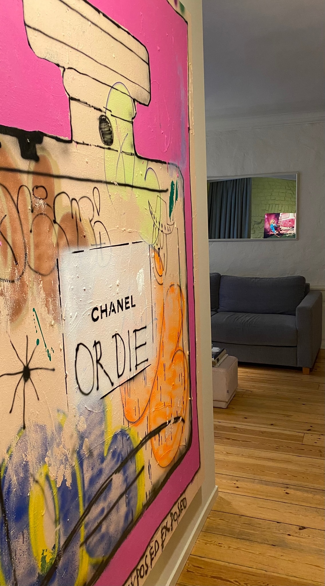 A vibrant mural depicting a Chanel perfume bottle is featured on a light-colored wall, adding a splash of color to the hallway. A grey sofa is visible in the adjacent room, with a glimpse of a decorative artwork on the opposite wall.