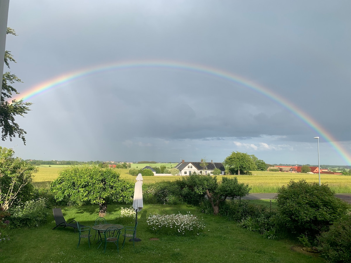 A vibrant rainbow arcs across a cloudy sky, providing a colorful contrast to the green fields below. In the foreground, a serene garden is visible, featuring areas of grass, flowers, and a few seating arrangements, inviting relaxation amidst the natural setting.