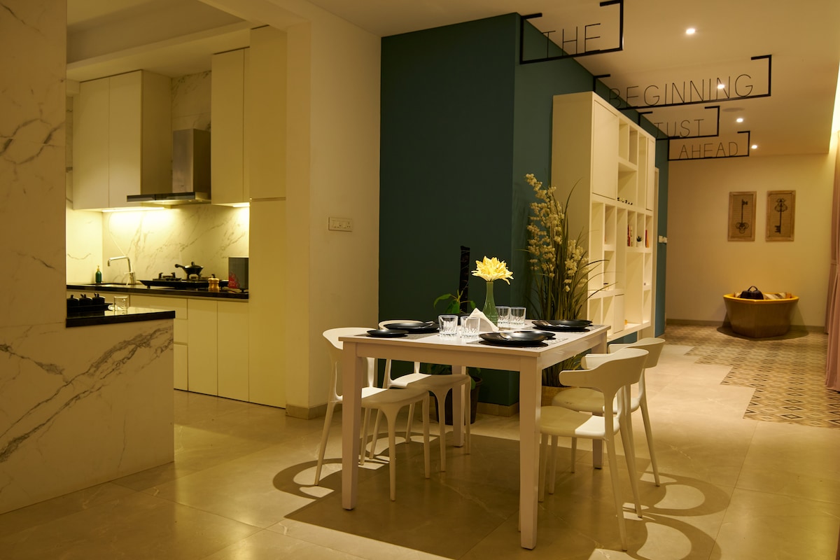 An inviting kitchen area is paired with a dining space featuring a white table and light-colored chairs. Soft lighting illuminates sleek cabinetry and a dark countertop. Decorative elements, including a potted plant and wall art, contribute to the room's modern design.