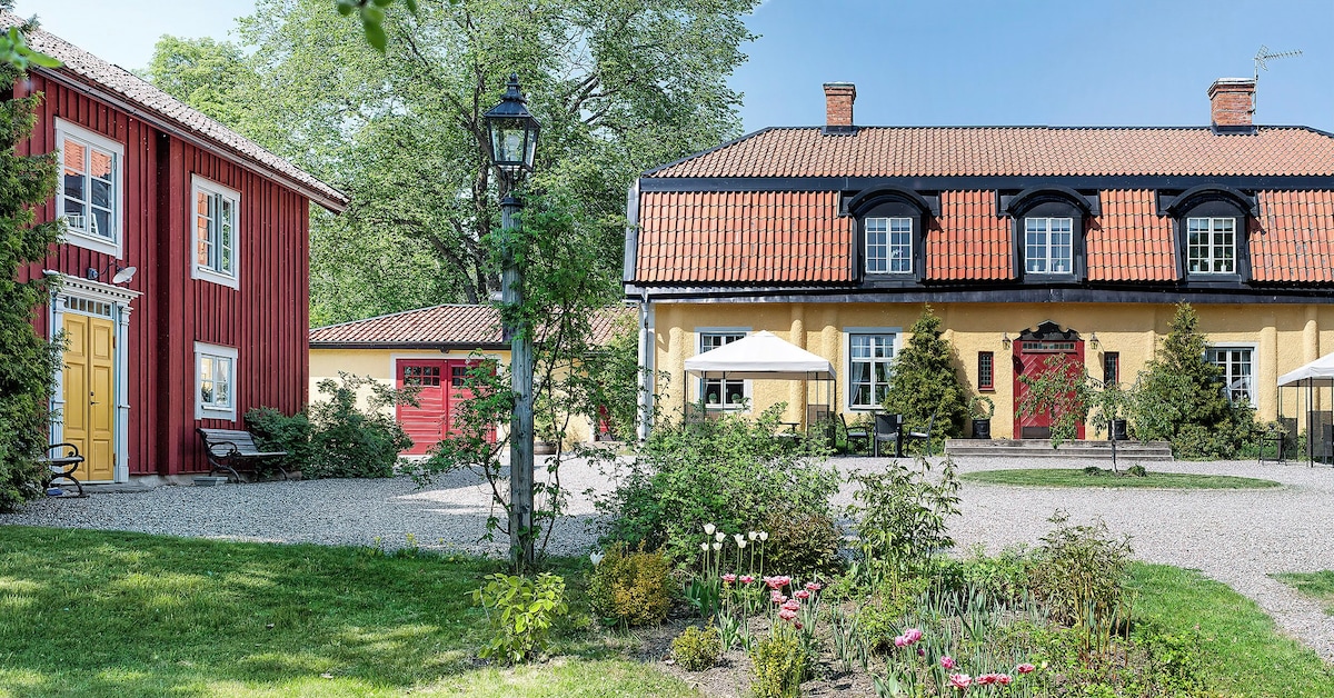 A charming historic estate is depicted, featuring a red wooden structure beside a yellow building with a black roof. Lush greenery surrounds the area, complemented by outdoor seating areas and umbrellas. Gravel pathways and blooming flowers enhance the serene outdoor ambiance.