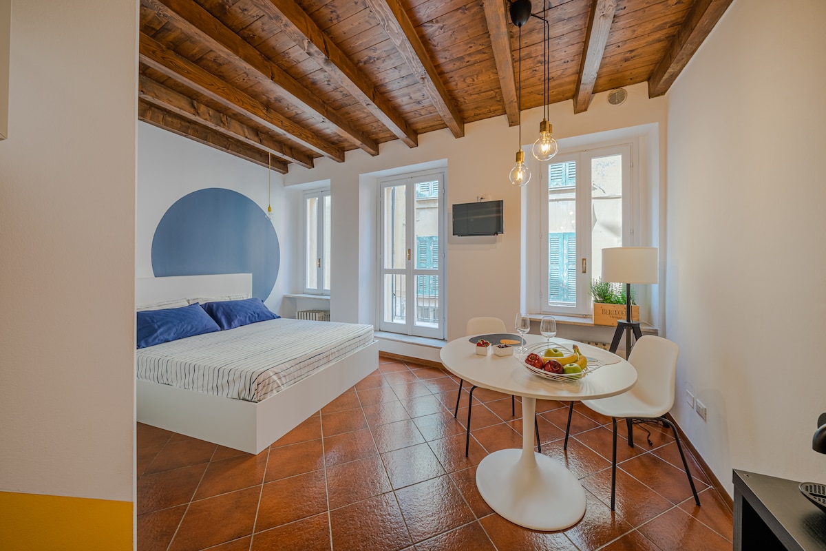A spacious room features a large bed with blue-accented pillows. Natural light enters through multiple windows, creating a bright atmosphere. A round dining table is set with breakfast items, and a flat-screen TV is mounted on the wall. Exposed wooden beams add character to the room.