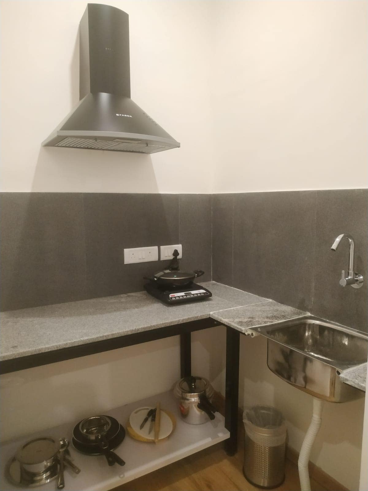A compact kitchenette is presented with sleek grey countertops. A range hood is mounted above a stovetop, with pantry essentials visible on the lower shelves. A stainless steel sink is situated beside the stovetop, complemented by a bin for waste disposal. Ample workspace is provided.