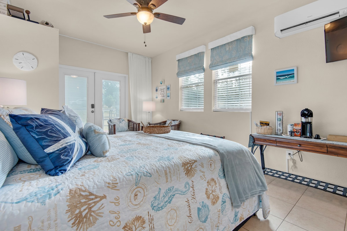 A spacious studio room includes a queen size bed adorned with ocean-inspired bedding. Natural light enters through two windows, highlighted by soft blue treatments. A comfortable seating area is nearby, along with a small desk featuring a coffee machine and decorative coastal artwork.
