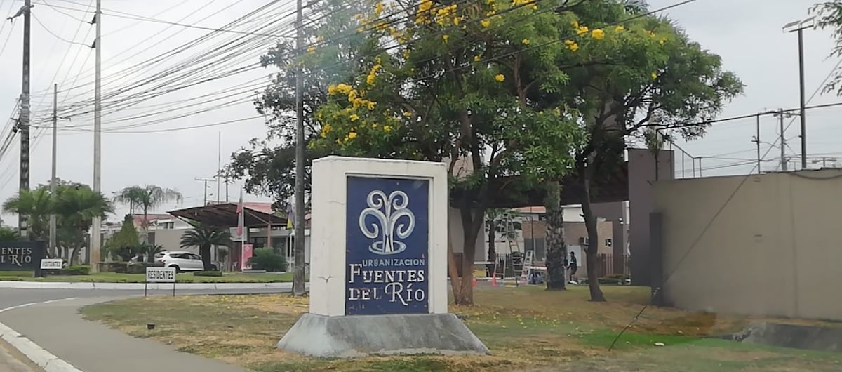 A community entrance sign is prominently displayed, featuring a decorative emblem and the name 'Fuentes del Río'. Surrounding greenery includes trees with yellow blooms, and a road leading into the neighborhood is visible, along with a glimpse of nearby utility poles.