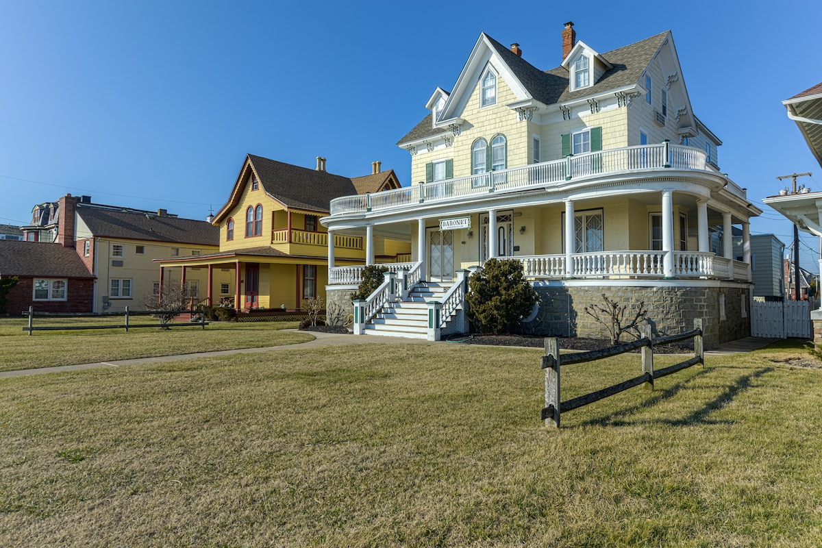 A grand historic mansion with a light yellow façade and white trim features a spacious front porch with multiple columns. Lush green grass surrounds the building, while neighboring structures with varying architectural styles are visible in the background under a clear blue sky.