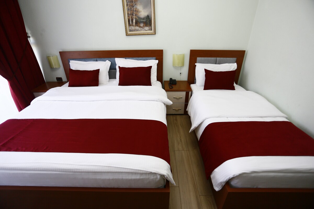 The room features two beds adorned with white linens and rich red accents. A nightstand is positioned between the beds, and a light fixture is mounted above each bedside. Natural light enters through a window covered with deep red curtains, enhancing the welcoming ambiance.
