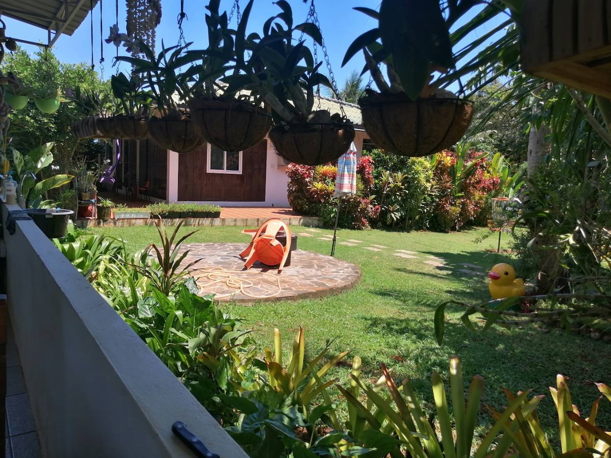 A lush garden area is visible, featuring well-maintained landscaping with a variety of plants and flowers. A circular stone area holds a bright orange chair, while hanging planters add depth overhead. A small decorative element resembling a duck can be seen on the nearby ground.
