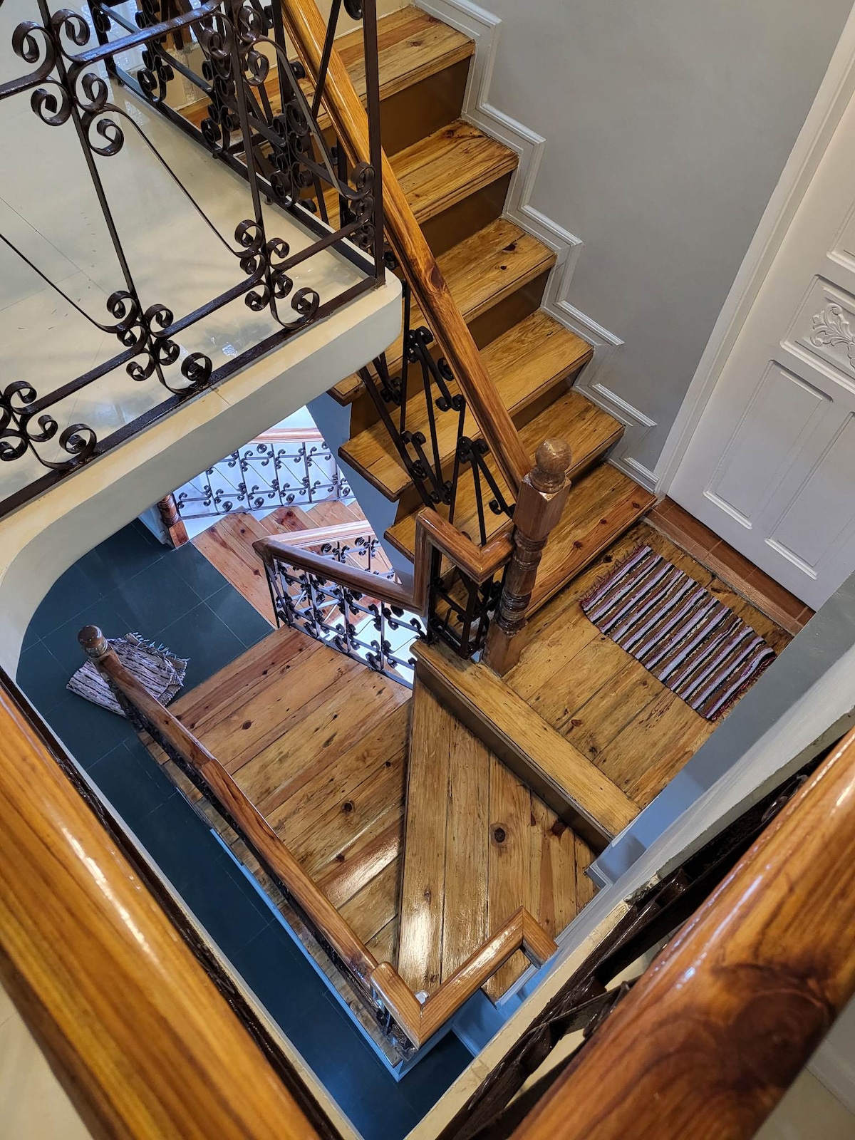 A spiral staircase made of polished wood is highlighted, featuring a wrought iron railing for safety. The wooden steps and floor are complemented by a small striped rug at the base. Natural light streams in from nearby windows, enhancing the warm tones of the wood.