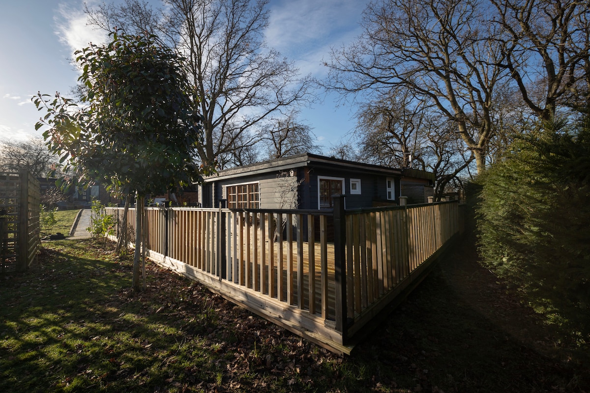 A wooden cabin is surrounded by trees, showcasing a spacious outdoor deck enclosed by a wooden railing. The cabin features large windows, allowing natural light to fill the interior. A well-maintained garden area is visible, enhancing the peaceful outdoor setting.
