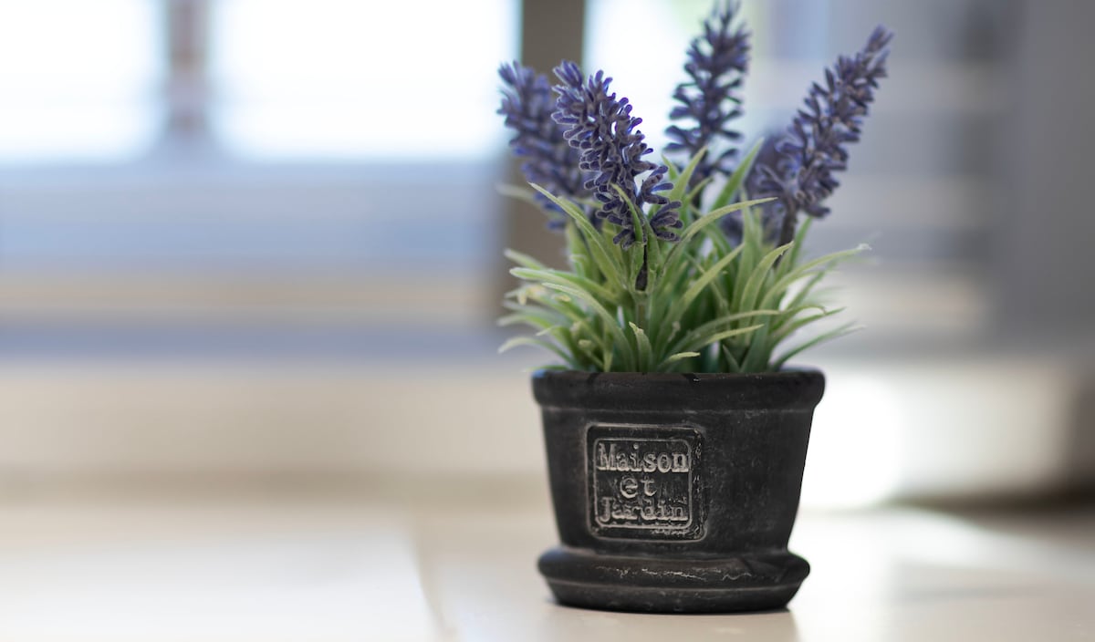A small black pot holds a cluster of faux lavender and greenery. The plant adds a touch of color to the setting, while light filters in softly from a nearby window.