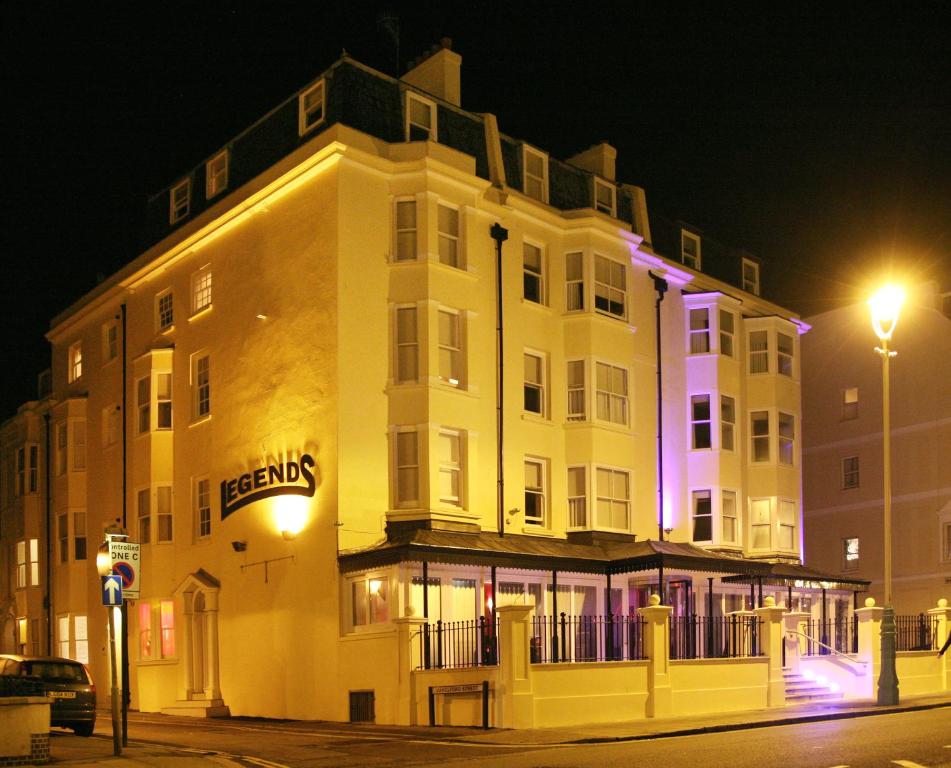 The exterior of the Legends bar and lodging is illuminated under the night sky, showcasing a three-story building with prominent signage. Soft lighting highlights the architectural features and outlines the balconies, while large windows reflect the ambient glow of the surroundings.