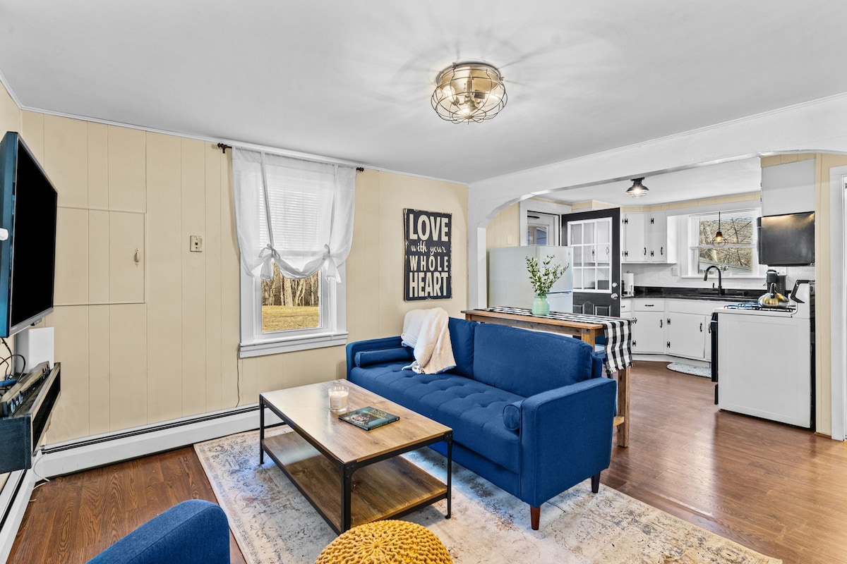 A bright living area features a blue sofa paired with a wooden coffee table and a simple rug. Light from windows illuminates the space, while a wall art piece with the message 'LOVE with your WHOLE HEART' enhances the atmosphere. The kitchen is visible in the background.