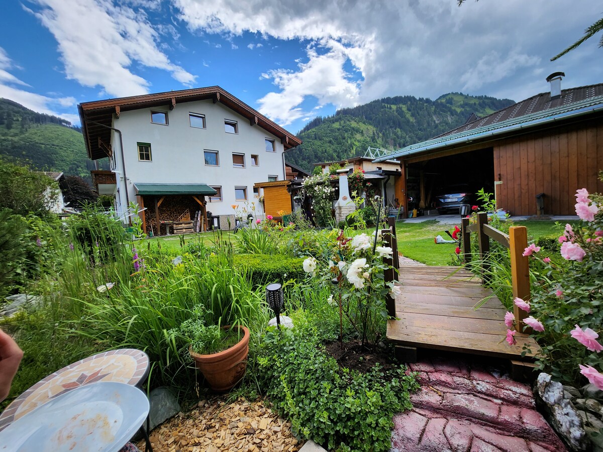 A spacious garden features a variety of lush greenery and blooming flowers. A wooden pathway leads through the landscaping, connecting to a building that showcases a balcony. The surrounding mountains provide a scenic backdrop under a partly cloudy sky.