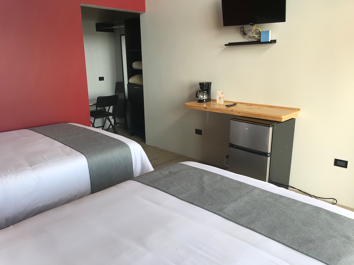 The room features two spacious beds with light grey accents on crisp white bedding. A wooden countertop displays a coffee maker and a mini refrigerator. The area is complemented by a mounted television and a closet with neatly stacked towels in the background.