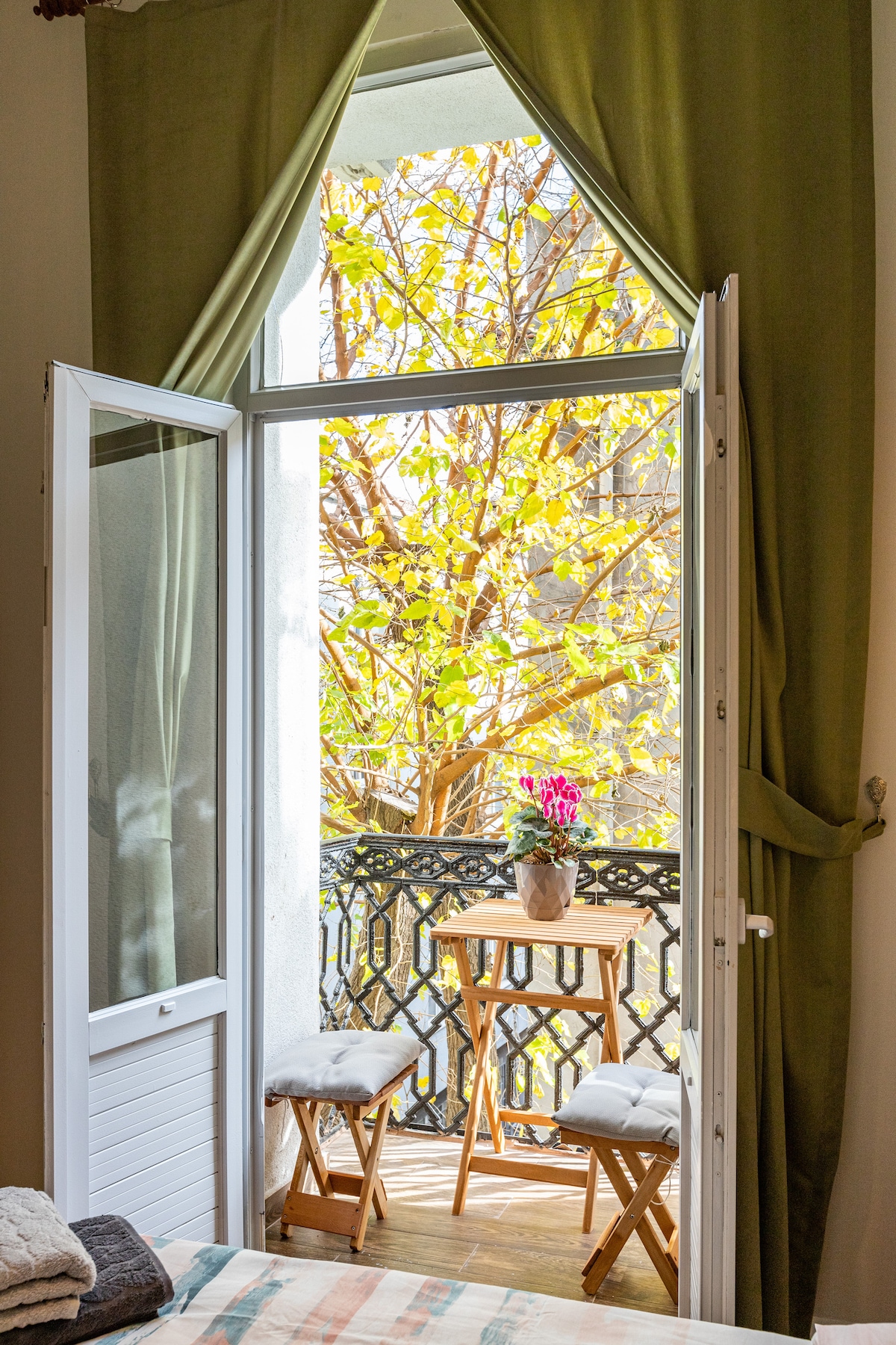 A balcony door opens to a view of vibrant autumn leaves, framed by a light green curtain. On the small balcony, a narrow table and two stools are positioned, while a potted plant adds a touch of color to the scene.