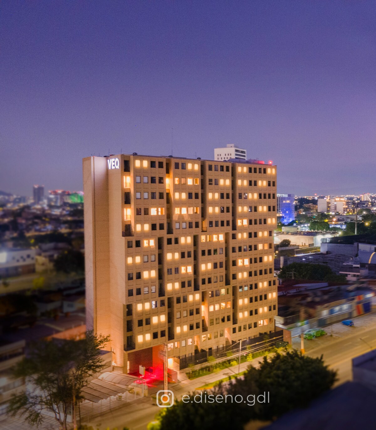 The building is illuminated at night, displaying multiple windows with warm lights. It features a modern design, standing tall in an urban setting. Surrounding elements include city lights in the distance, creating a lively backdrop.