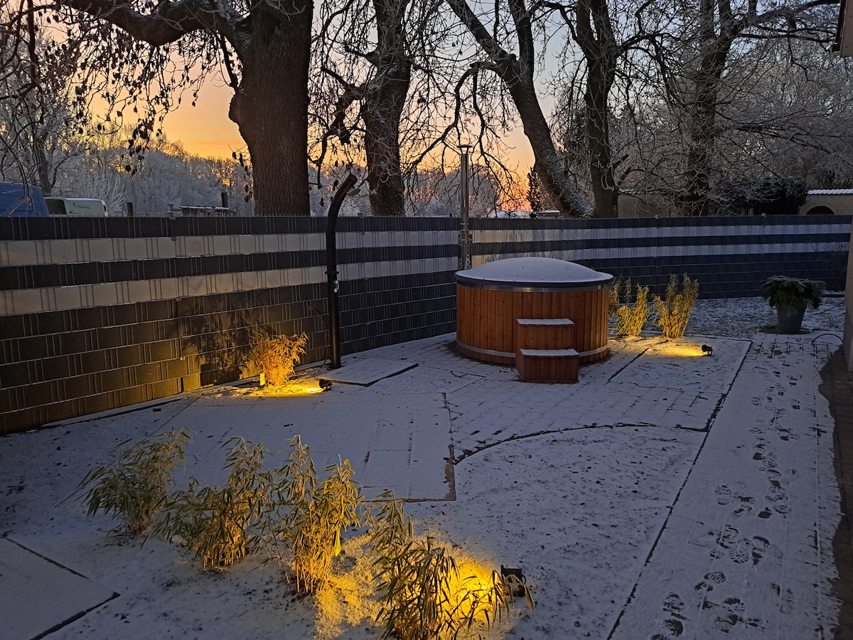 A snow-covered garden features a wooden hot tub surrounded by soft, warm lighting. The path is lined with landscaped greenery, and a fence provides privacy. Trees are visible against a colorful sky as the day begins to unfold.