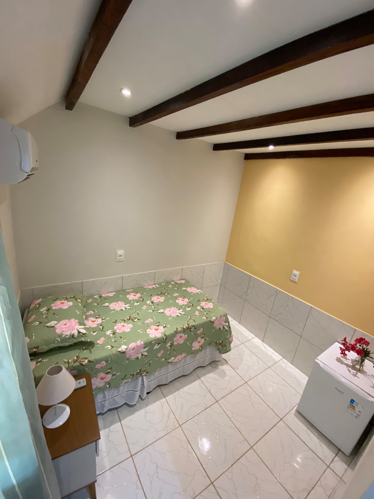 A compact room features a floral-patterned bedspread over a spacious bed, with light-colored walls and wooden beams overhead. A small refrigerator is positioned beside a bedside table, illuminated by soft lighting. A window draped with sheer curtains allows natural light to brighten the space.