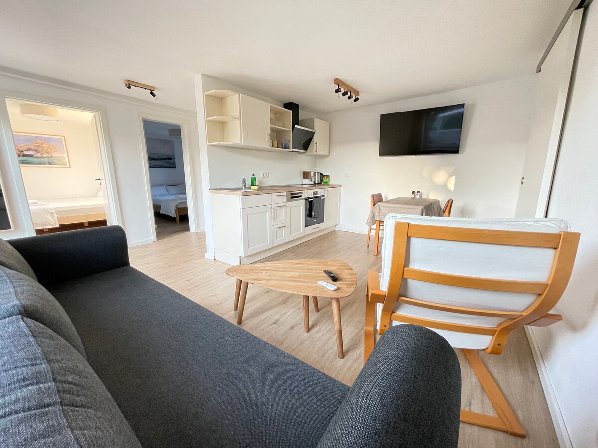 A combined living area features a modern kitchen with light cabinetry and a flat-screen TV mounted on the wall. A cozy sofa and wooden coffee table are present, along with two simple armchairs. Natural light fills the space, creating an inviting atmosphere.