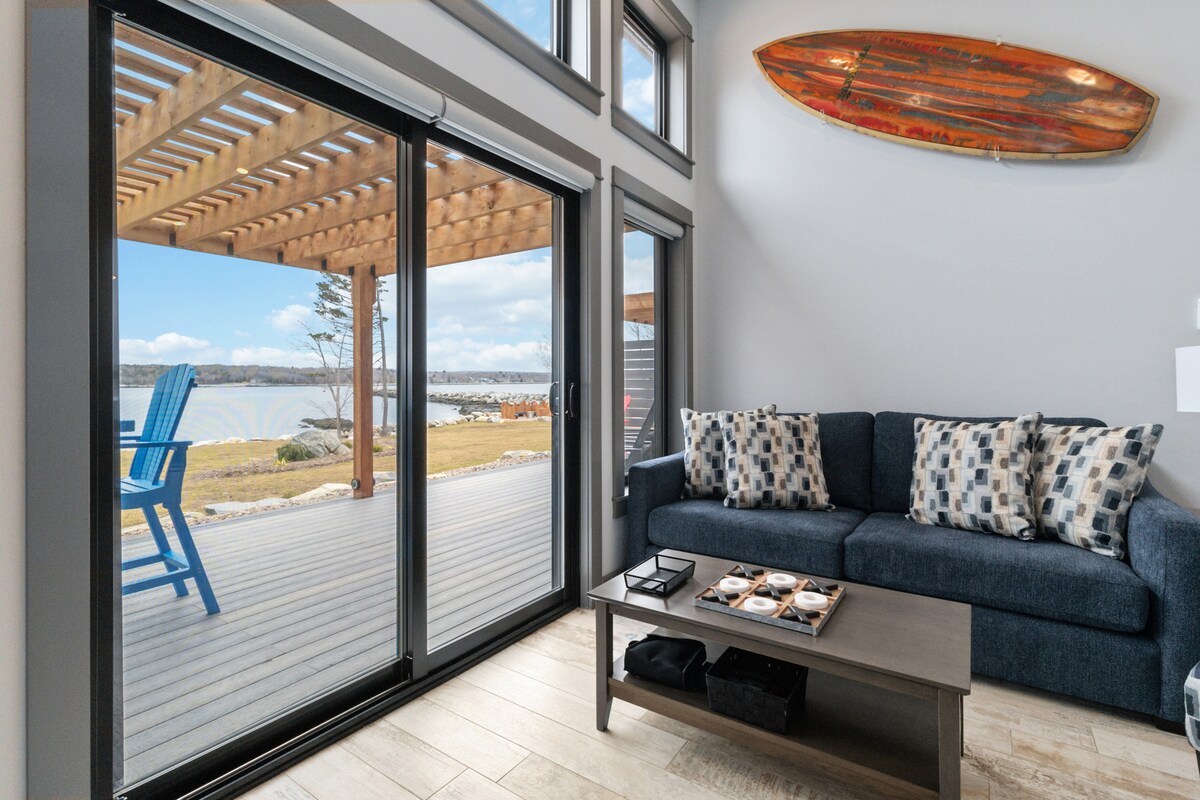 A comfortable living area features large glass doors that open to a spacious wooden deck overlooking the water. A dark blue sofa adorned with patterned cushions is positioned near a coffee table displaying decorative items. Natural light fills the space, highlighting the contemporary design.