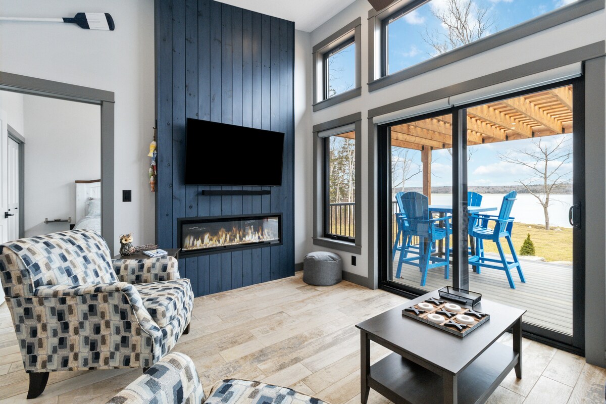 A light-filled living area features large glass doors opening to a deck overlooking the water. Comfortable patterned armchairs are positioned near a modern fireplace set within a blue accent wall. An understated coffee table sits centrally, enhancing the functional space.