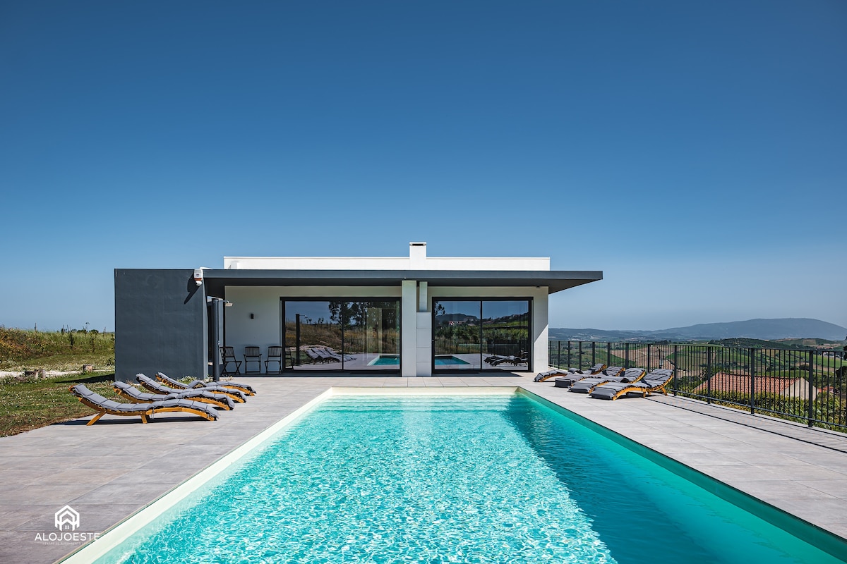 A modern house with large glass doors opens onto a spacious terrace and a sparkling pool. Sun loungers are positioned around the pool, offering views of the surrounding landscape. The clear blue sky enhances the tranquil atmosphere of the outdoor space.